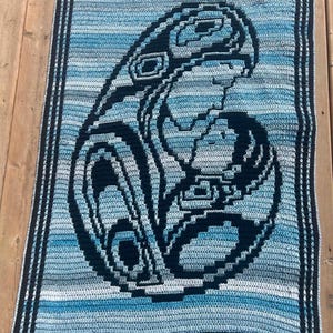 May include: A rectangular crochet blanket featuring a dark navy blue graphic design of a parent and child in a stylized form. The background consists of horizontal stripes in varying shades of light blue, teal, and white, framed by a dark navy border.