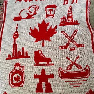 May include: A white crocheted blanket with a red border featuring various Canadian-themed icons in red, including a maple leaf, a moose, mountains, a beaver, a coffee cup, a lighthouse, the CN Tower, hockey sticks, a skate, a syrup jug, and an inukshuk.