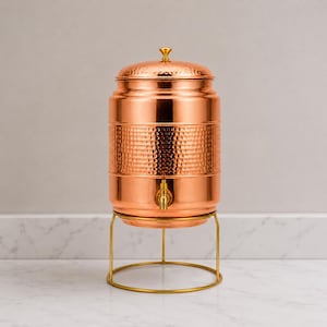 May include: A cylindrical copper water dispenser featuring a hammered texture on the central section, a removable lid with a small gold-toned knob, and a gold-toned spigot. The vessel rests on a thin, circular gold-toned metal wire stand.