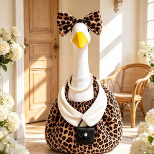 May include: A white goose figurine wearing a leopard print bow on its head, a white pearl necklace, a white collared garment, and a leopard print body covering with a small black purse featuring a single white pearl accent attached to the front.