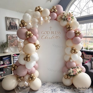 Op de afbeelding: Een witte boogvormige achtergrond met de tekst GOD BLESS Charlotte en een kruis, omringd door een ballonslinger in tinten crème, oudroze en metallic goud, geaccentueerd met doorzichtige confettiballonnen en bloemstukken.