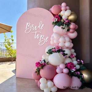 Op de afbeelding: Een roze boogvormige achtergrond met de witte tekst Bride to be, versierd met een grote ballonslinger in tinten roze, wit en goud, geaccentueerd met kunstmatige roze bloemen en groene bladeren voor een feestelijke decoratie.
