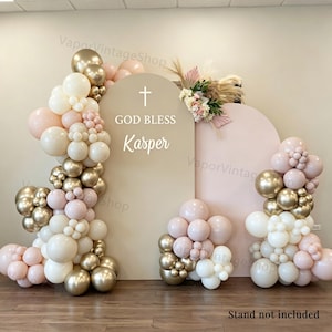 Puede incluir: Un conjunto de fondos arqueados en beige y rosa claro con un icono de cruz blanca y el texto "GOD BLESS Kasper" en blanco. La decoración incluye una gran guirnalda de globos en tonos crema, rosa suave y oro metálico, junto con un arreglo floral.