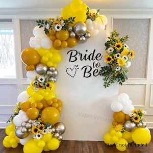 Op de afbeelding: Een witte boogvormige achtergrond met de zwarte tekst Bride to Be en een hartje, versierd met een grote ballonslinger in geel, wit en metallic zilver, geaccentueerd met kunstmatige zonnebloemen en groene bladeren als decoratie.
