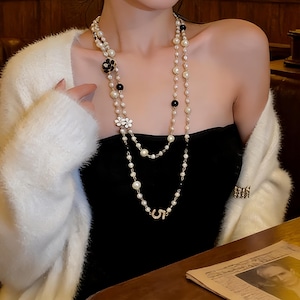 May include: A long layered necklace featuring white faux pearls, small black beads, and decorative accents including a black flower, two white flowers, and a metallic number five charm. The necklace is worn over a black strapless top and a white cardigan.