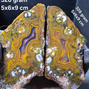 Può includere: Due metà di una pietra d'agata lucidata con bande concentriche gialle, arancioni, marroni e bianche con centri traslucidi. Il testo sull'immagine recita 328 gram e 5x6x9 cm. La pietra mostra intricati motivi minerali e inclusioni cristalline.