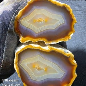 May include: Two polished agate slices featuring concentric bands of amber, yellow, and white, with a central crystalline cavity. The text 518 geam 5x6x12 cm and Galaxy S25 Ultra is visible. The stones display a translucent quality and natural mineral patterns.