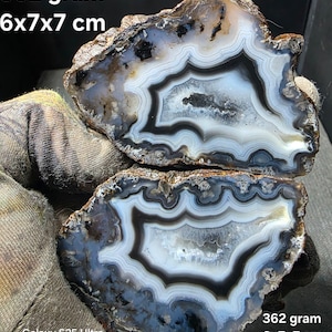 Puede incluir: Una rodaja de geoda de ágata pulida con bandas concéntricas de color blanco, gris y negro, con un centro cristalino. El espécimen pesa 362 gramos y mide 6x7x7 cm. La piedra muestra formaciones minerales naturales y bordes exteriores rugosos.