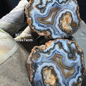 Puede incluir: Dos rodajas de ágata pulida con bandas concéntricas de color blanco, gris y marrón translúcido con inclusiones minerales de color óxido. El texto 324 gram 5x6x7cm es visible. Las piedras muestran patrones naturales intrincados.