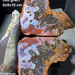 Puede incluir: Dos rodajas de ágata pulida con bandas intrincadas en tonos naranja, blanco y marrón con inclusiones cristalinas oscuras. El texto 988 gram 6x9x10 cm es visible en la imagen. La piedra muestra un patrón complejo de anillos concéntricos.