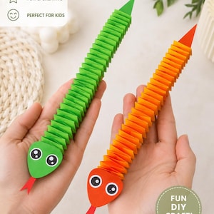 Può includere: Due serpenti di carta con corpi piegati a fisarmonica in verde brillante e arancione. Ogni serpente ha una testa di carta piatta con grandi occhi bianchi e neri e una lingua rossa appuntita. Il serpente verde ha la punta della coda verde e quello arancione arancione.