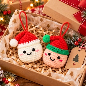 Puede incluir: Dos adornos redondos de ganchillo con caras sonrientes y mejillas rosadas. Uno es blanco con un gorro de Papá Noel rojo y pompón blanco, el otro es rosa claro con un gorro de elfo rojo y verde y un pompón verde. Ambos tienen lazos rojos.