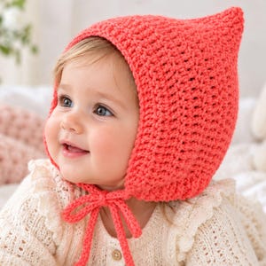 May include: A bright coral-coloured crochet bonnet featuring a pointed top and matching chin ties. The headwear is crafted with a textured yarn stitch pattern, providing a snug fit around the head and ears with a secure bow tied under the chin.