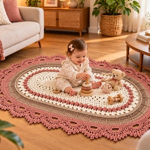 May include: An oval-shaped crochet rug featuring a central cream-coloured section, a middle band of light brown, and a wide, decorative scalloped border in dusty rose. The rug has a textured, intricate pattern with small raised bobble details.