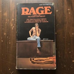 Op de afbeelding: Een paperback boek getiteld RAGE door Richard Bachman met een zwarte kaft. De kaft toont een persoon die op een bureau in een klaslokaal zit, met de tekst: HIS TWISTED MIND TURNED A QUIET CLASSROOM INTO A DANGEROUS WORLD OF TERROR. Benen zijn zichtbaar.