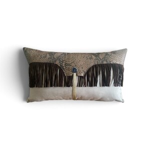 May include: A rectangular throw pillow featuring a top panel with a tan and brown snakeskin pattern, a middle section with dark brown fringe, and a solid light beige bottom panel. A thin gold cord hangs from a small blue circular button.