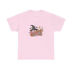 May include: A light pink short-sleeve t-shirt featuring a graphic print with the text "BASIC WITCH" in retro-style lettering. The design includes a black witch hat with a spiderweb pattern, a coffee cup with a smiley face, bats, and a lightning bolt.