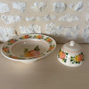 Może przedstawiać: Kremowa ceramiczna maselniczka składająca się z szerokiej, płytkiej miseczki i pasującej wypukłej pokrywki. Oba elementy ozdobione są ręcznie malowanym wzorem pomarańczowych dyń, zielonych liści i pnączy z delikatnym obrzeżem w kolorze pomarańczowym.