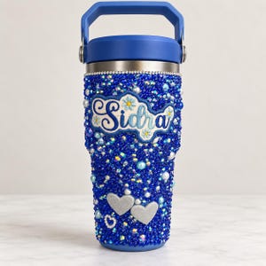 Puede incluir: Un vaso térmico azul con asa, cubierto completamente de pedrería y perlas azules. El frente presenta un parche bordado con el nombre Sidra en letra cursiva azul, decorado con pequeñas flores y dos adornos plateados en forma de corazón.