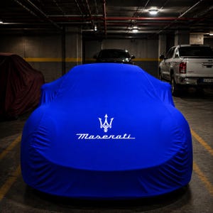 May include: A vibrant blue car cover draped over a vehicle, featuring the white Maserati trident logo and the brand name printed clearly in the centre. The fabric is smooth and form-fitting, highlighting the contours of the car underneath.