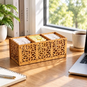 Könnte beinhalten: Ein rechteckiger Teebeutel-Organizer aus Holz mit einem lasergeschnittenen Blumenmuster an der Vorderseite und den Seiten. Die Box ist in drei Fächer unterteilt, die mit Teebeuteln in weißen und hellgelben Papierverpackungen gefüllt sind.