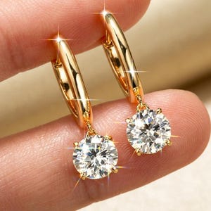 May include: A pair of gold hoop earrings, each featuring a single round, brilliant-cut clear gemstone suspended from the bottom. The metal has a polished, reflective finish, and the stones exhibit multiple facets that catch and reflect light.