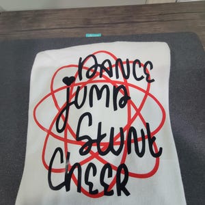May include: A white t-shirt featuring a red graphic of overlapping elliptical shapes resembling an atom. Overlaid on the red graphic is black text that reads DANCE, JUMP, STUNT, and CHEER, with a small black heart icon above the word JUMP.