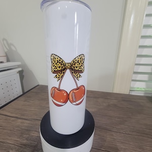 Op de afbeelding: Een witte cilindrische beker met een doorzichtig deksel, voorzien van een afbeelding van twee oranje footballs in de vorm van kersen, verbonden door steeltjes aan een grote strik met luipaardprint. Het ontwerp bevat witte accenten.