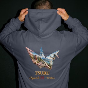 Op de afbeelding: Een leigrijze hoodie met een grote afbeelding op de achterkant van een origami kraanvogel. Het ontwerp van de kraanvogel bevat een landschap met een pagode, kersenbloesems en een berg. De tekst luidt TSURU, Origami-bu en 一枚の紙から.