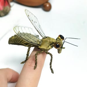 Può includere: Una statuetta di insetto realizzata a mano con fibre vegetali verdi intrecciate. L'insetto presenta ali traslucide venate, due prominenti occhi sferici marrone scuro e sottili antenne in filo metallico nero. Corpo segmentato.