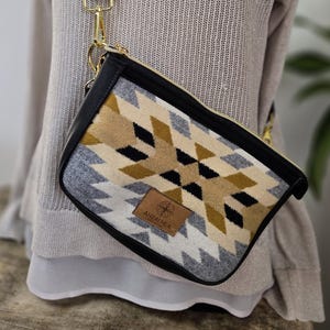 May include: A crossbody bag with a geometric pattern in black, tan, and grey. The bag has a black leather trim and a black adjustable strap with gold-coloured hardware. A small leather tag with a logo is attached to the front.