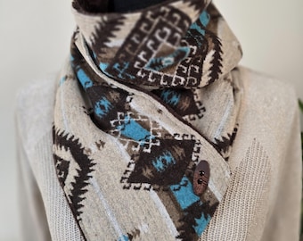 Puede incluir: Bufanda estampada en beige y marrón, con detalles turquesa y cierre de botón marrón. La bufanda se muestra en un maniquí, destacando su diseño y textura. El patrón incluye formas y líneas geométricas.