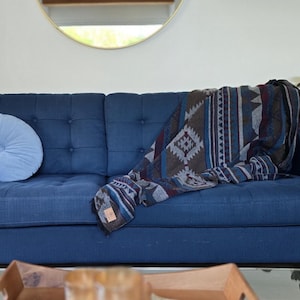 Może przedstawiać: Granatowa sofa z dwiema jasnoniebieskimi okrągłymi poduszkami i wzorzystym kocem. Koc ma geometryczne wzory w odcieniach niebieskiego, brązowego i szarego. Złote lustro wisi na ścianie nad sofą.