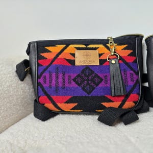 May include: A patterned crossbody bag in vibrant colours: orange, red, purple, and black. It features black leather trim, a gold zip, and a leather tassel. The brand name "AHZALHEA" is displayed on a leather patch.