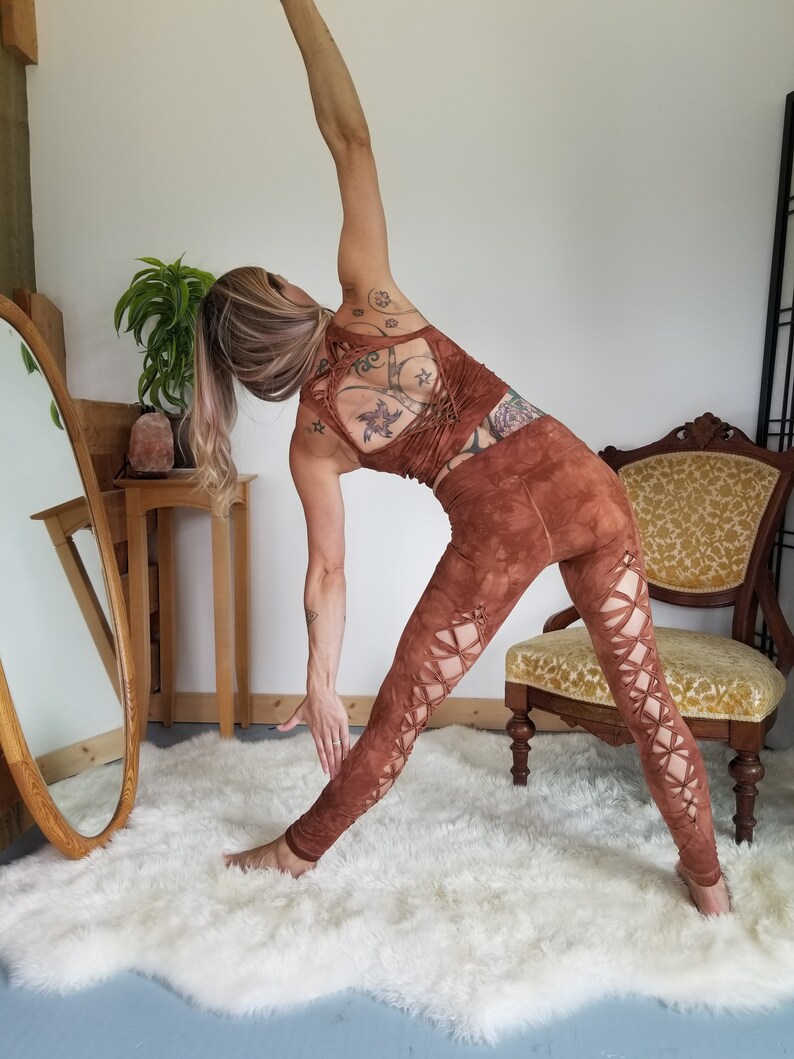 Pode incluir: Uma mulher vestindo leggings marrons com recortes e um top marrom combinando com as costas cruzadas. Ela est&aacute; em p&eacute; em um tapete branco e felpudo e est&aacute; esticando o bra&ccedil;o acima da cabe&ccedil;a.