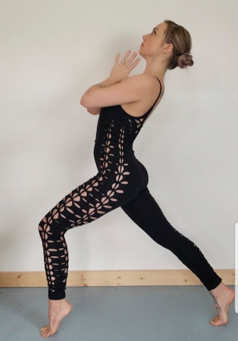 May include: A woman wearing a black one-piece outfit with a cut-out floral design. The outfit is form-fitting and has a high neckline. The woman is standing in a yoga pose with her arms raised above her head.