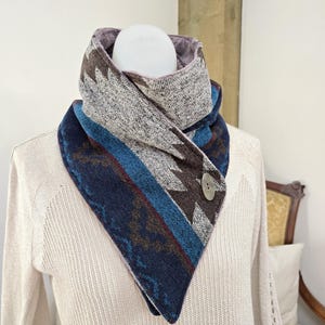 May include: A triangular scarf with a button closure. The scarf features a geometric pattern in shades of blue, brown, and gray. The scarf is displayed on a mannequin, showcasing its design and texture. The scarf is a cozy accessory.