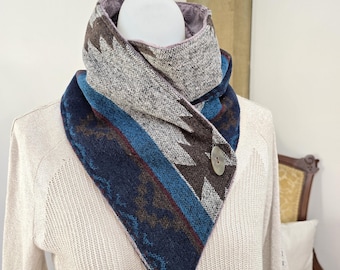 Puede incluir: Bufanda triangular con cierre de botón. Presenta un patrón geométrico en tonos azules, marrones y grises. Se muestra en un maniquí, resaltando su diseño y textura. Un accesorio acogedor para el invierno.