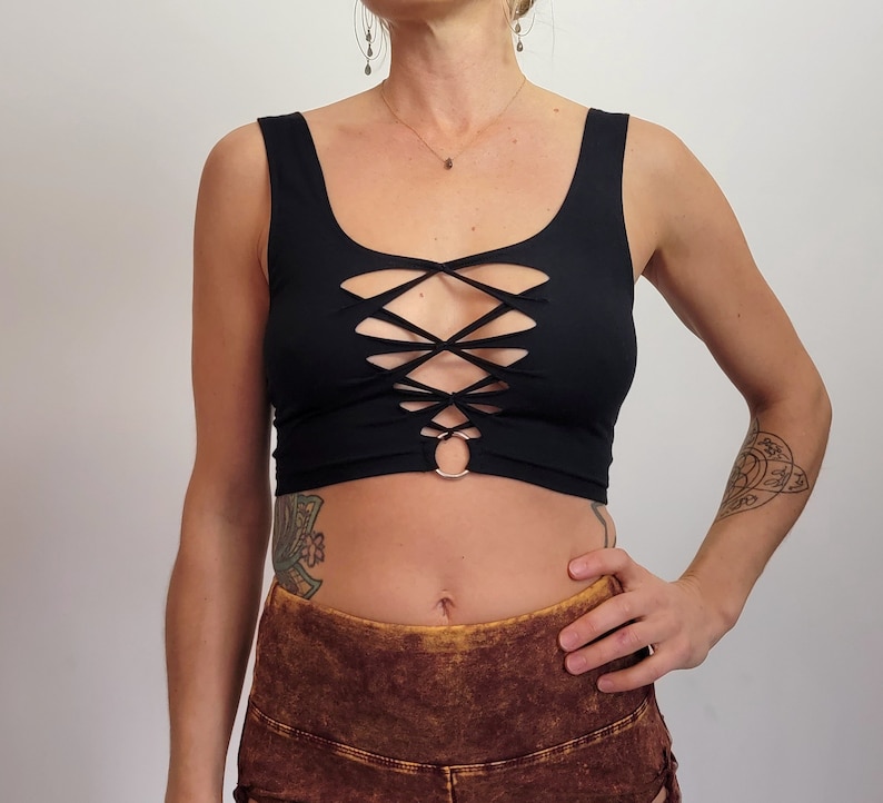 May include: Black crop top with a criss-cross lace-up design in the front. The top has a scoop neckline and a ring detail at the center.