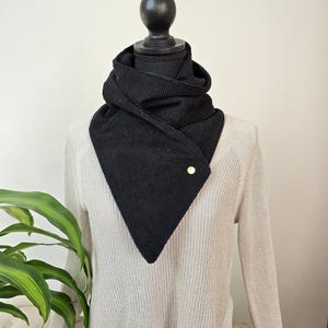 May include: A black corduroy scarf with a triangular design, fastened with a gold-colored button. The scarf is draped over a light-colored sweater on a mannequin. A green plant is visible in the background.