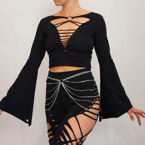 May include: Black long-sleeved crop top with a lace-up detail in the front and bell sleeves. The top is paired with black pants with cutouts and a silver chain belt.