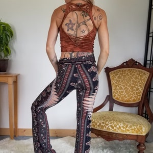 Pode incluir: Cal&ccedil;as de boca de sino com padr&atilde;o preto e vermelho e fendas nas laterais. As cal&ccedil;as s&atilde;o usadas por uma pessoa com um top curto marrom e tatuagens.