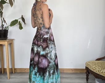 Può includere: Un abito lungo stile halter con un motivo tie-dye nei toni del viola, marrone e turchese. L'abito ha una gonna fluida e un collo halter. La modella indossa l'abito in una stanza con un muro bianco.