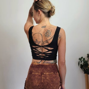 May include: Black crop top with a criss-cross back design. The top is made of a stretchy, comfortable fabric and has a relaxed fit. The model is wearing brown shorts with a distressed look.