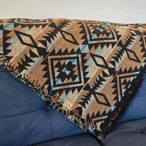 Puede incluir: Una manta tejida con un patrón geométrico en tonos marrón, negro y turquesa. La manta tiene un borde con flecos y está colocada sobre un sofá azul. El diseño presenta formas de diamantes y zigzags.