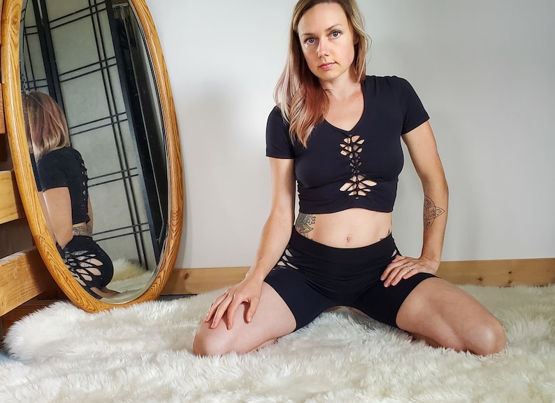 May include: A woman wearing a black crop top with a lace-up design and black shorts with cutouts. She is kneeling on a white fluffy rug.