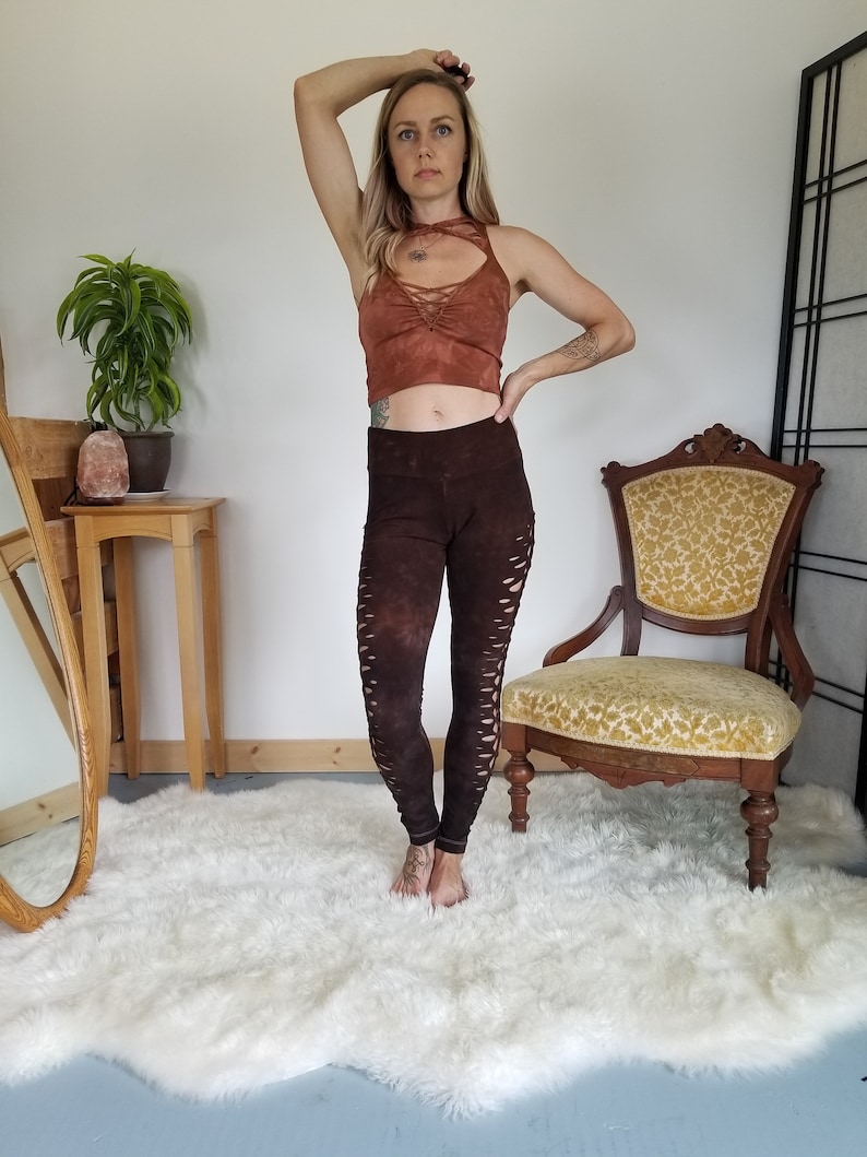 Pode incluir: Uma mulher vestindo um top marrom com um detalhe de renda e leggings marrons com recortes. Ela est&aacute; em p&eacute; em um tapete branco e felpudo na frente de uma cadeira de madeira.