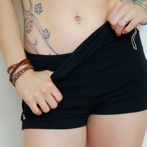 May include: A woman wearing black shorts with side cutouts and a floral tattoo on her hip. She is wearing multiple bracelets on her wrist and a belly button ring.