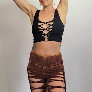 May include: A black crop top with a criss-cross design and a metal ring at the center. The top is worn with brown pants that have a distressed look and multiple cutouts.