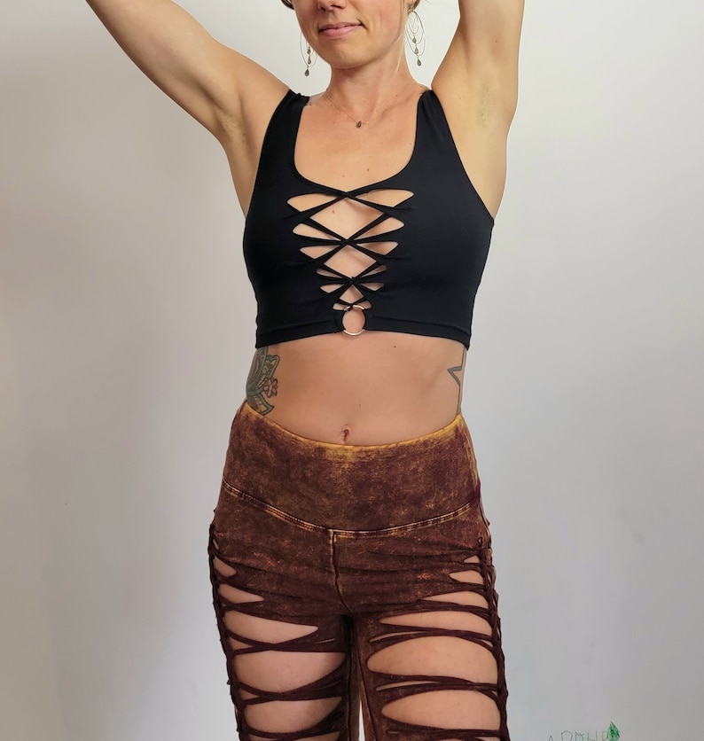 May include: A black crop top with a criss-cross design and a metal ring at the center. The top is worn with brown pants that have a distressed look and multiple cutouts.